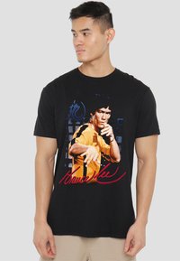 Black t-shirt featuring a colorful print of a male figure in a yellow outfit. Includes red text "Bruce Lee" and blue design elements.