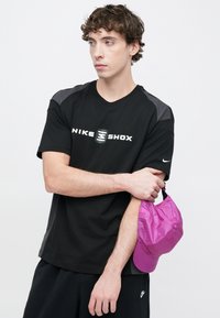 Μαύρο κοντομάνικο μπλουζάκι με γκρι λεπτομέρειες, με λευκό λογότυπο "NIKE SHOX". Κρατώντας ένα ροζ, ελαφρύ καπέλο με στρογγυλή κουμπί λεπτομέρεια.