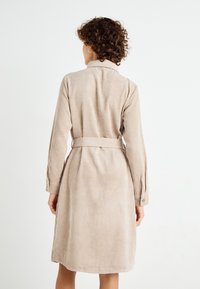 Beige kordflor klänning med bälte i midjan, långa ärmar och vertikal ribbad textur. Har en krage och knäppta manschetter.