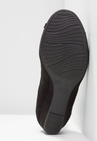 Semelle de chaussure noire avec motif ondulé et petit talon appuyée contre un coin de mur blanc, taille marquée 37H sur la semelle.