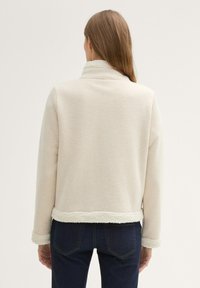 Veste en polaire beige avec un col et un ourlet doux et texturé. Présente des manches longues et une silhouette décontractée. Un jean en denim foncé complète le look.