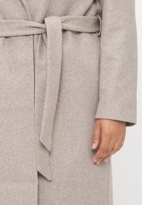 Cappotto in lana grigio chiaro con una finitura testurizzata, caratterizzato da una cintura annodata in vita e maniche larghe, ripreso dalla sezione centrale con una mano visibile.
