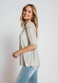 Hellbeige, lockere Bluse mit 3/4-Ärmeln und rohen Kanten. Der Stoff wirkt weich mit einer leicht strukturierten Oberfläche.