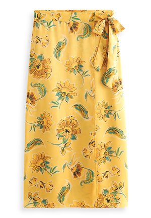 Femme portant une robe midi jaune à fleurs avec manches bouffantes et ceinture à la taille, associée à des sandales dorées à talons hauts, debout près d’un mur blanc.