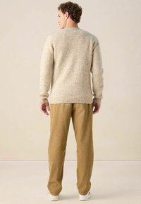 Mann, der einen hellbeige gesprenkelten Strickpullover und eine sandfarbene Cordhose trägt, steht mit dem Rücken zur Kamera vor einem schlichten Hintergrund.