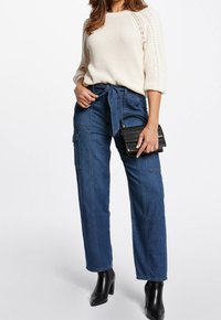 Beige gestrickter Pullover mit lockeren Ärmeln, kombiniert mit hoch taillierten blauen Jeans-Cargohosen mit einem Bindegürtel. Schwarze Handtasche und Ankle-Boots.