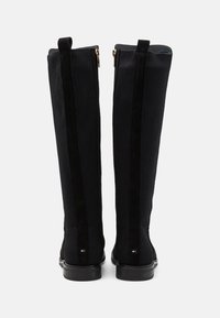Black knee-high boots feature a ribbed material with suede accents, gold zippers, and a flat sole. Both sides display a discreet logo detail.
