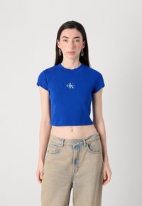 Une jeune femme se tient devant un fond gris clair, portant un crop top bleu ajusté avec un logo Calvin Klein et un jean délavé.