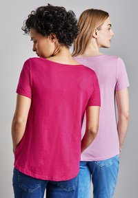 Dos mujeres que aparecen de espaldas, vestidas con camisetas casuales; una es de color rosa brillante y la otra es de color púrpura claro. Las camisetas tienen mangas cortas y un escote redondo.