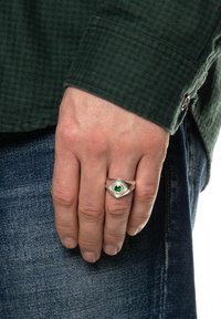 Anel sigiloso prateado com uma pedra verde, apresentando uma forma hexagonal e detalhes gravados. Usado numa mão que repousa contra um tecido de ganga.