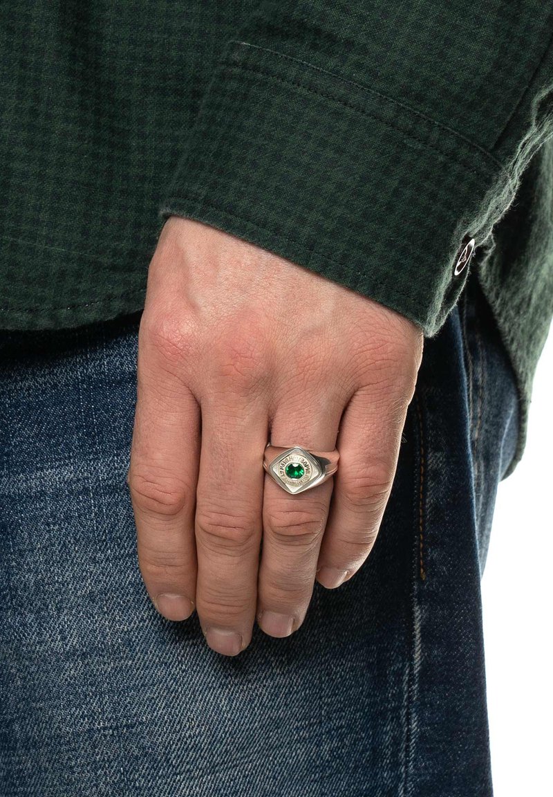 Anel sigiloso prateado com uma pedra verde, apresentando uma forma hexagonal e detalhes gravados. Usado numa mão que repousa contra um tecido de ganga.