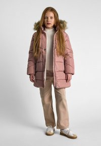 Manteau rose en duvet avec une capuche doublée de fourrure, doté d'une fermeture éclair à l'avant et de boutons-pression, associé à un pantalon beige et des baskets blanches.