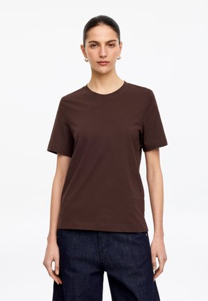 Femme aux cheveux foncés attachés, portant des boucles d’oreilles créoles en or, un t-shirt marron uni à manches courtes et un jean bleu foncé, debout sur un fond blanc.