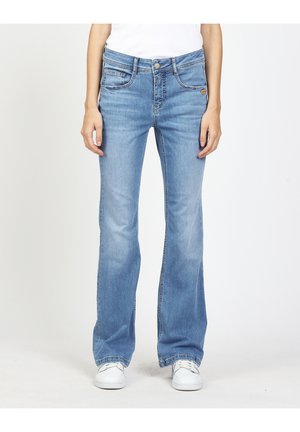 Person trägt hellblaue Bootcut-Jeans und weiße Sneaker, steht vor einem schlichten weißen Hintergrund.