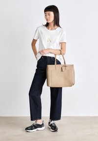 Sac fourre-tout beige à forme structurée avec poignées supérieures, associé à un t-shirt blanc arborant un logo doré et un jean large bleu foncé.