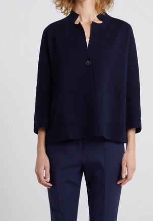Frau trägt eine marineblaue, langärmlige jacke mit knöpfen, zwei Vordertaschen und passende marineblaue Hose.