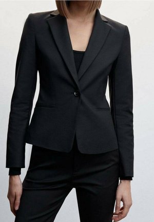 Vrouw draagt een getailleerde zwarte blazer met een enkele knoop en zwarte broek tegen een neutrale achtergrond.