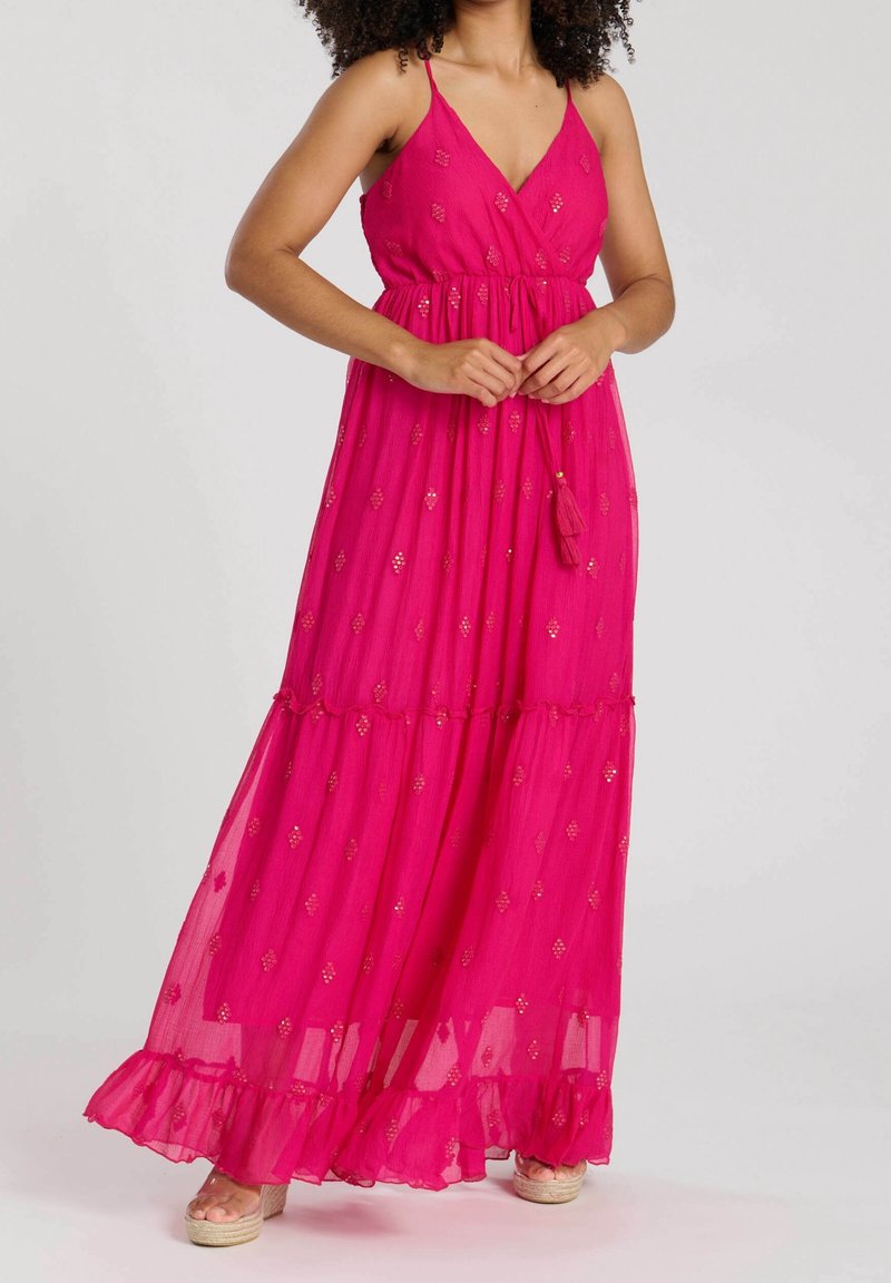 Femme portant une robe longue rose vif sans manches avec des accents en sequins dorés et un ourlet à volants, debout, les mains jointes à la taille.