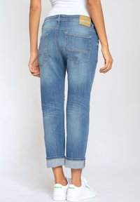 Vista trasera de una persona que lleva puestos unos jeans azules arremangados y unas zapatillas Reebok blancas contra un fondo claro y liso.