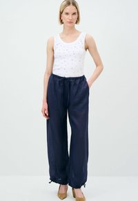 Witte mouwloze top met kleine decoratieve studs, gecombineerd met losvallende donkerblauwe broek met een elastische tailleband en manchetten.