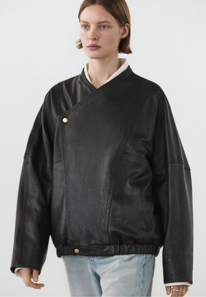 Giovane con capelli corti che indossa una giacca di pelle nera larga con parte frontale asimmetrica e jeans azzurri chiari su uno sfondo semplice.