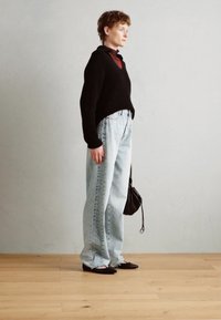 Suéter de punto negro con escote en V, corte holgado; jeans de tiro alto azul claro con costura lateral; bolso negro, zapatos planos negros; fondo neutro.