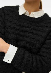 Svart stickad tröja med en texturerad vågmönster, rundad halsringning och sänkta axlar. En vit skjorta med krage syns under.