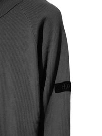 Close-up of a gray ribbed knit sweater sleeve with a black patch featuring partially visible white text on a white background.