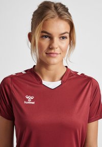 Bordeaux sportshirt met korte mouwen, V-halsontwerp en witte accenten op de schouders. Bevat logo op de voorkant. Gladde textuur.