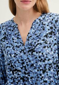 Blue patterned blouse featuring a v-neck, button detail, and pleats, made from lightweight fabric with a mix of black and light blue spots.