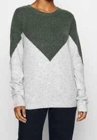 Maglione a maglia grigio e verde scuro con motivo a zig zag, scollatura rotonda e maniche lunghe con polsini a coste. Texture morbida.