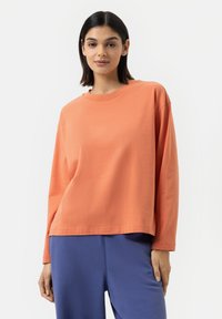 T-shirt à manches longues, col rond, en coton corail-orange. Coupe décontractée, texture lisse, avec des détails de couture subtils à l'ourlet. Associé à un pantalon bleu.