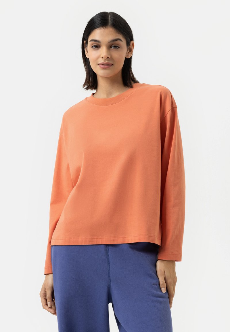 T-shirt à manches longues, col rond, en coton corail-orange. Coupe décontractée, texture lisse, avec des détails de couture subtils à l'ourlet. Associé à un pantalon bleu.