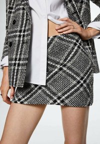 Jupe mini à carreaux en tissu texturé noir et blanc, associée à une chemise blanche et un blazer à carreaux noirs. Détail de la main visible sur la hanche.