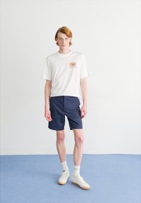 T-shirt bianca a maniche corte con logo grafico, abbinata a pantaloni corti blu navy. Il modello indossa sneakers bianche con accenti verdi e calze bianche.