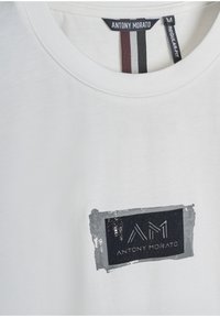 Camiseta blanca de algodón con cuello redondo, que presenta un parche rectangular gris con texto negro y un borde texturizado.