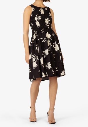 Kvinde iført en ærmeløs sort A-formet kjole med hvidt blomsterprint, smocket liv, lommer og nøgenfarvede hæle med ankelrem.