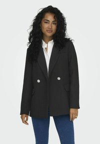 Blazer noir texturé avec un design croisé, deux poches avant et des boutons décoratifs en forme de fleur, porté sur une chemise blanche et un jean.