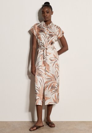 Femme portant une robe midi boutonnée à manches courtes, ceinturée, avec un motif abstrait de feuilles marron et beige, et des sandales plates marron.