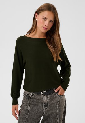 Femme aux longs cheveux bruns porte un haut vert foncé à manches longues et un jean gris délavé taille haute avec une ceinture noire, posant avec une main dans la poche.