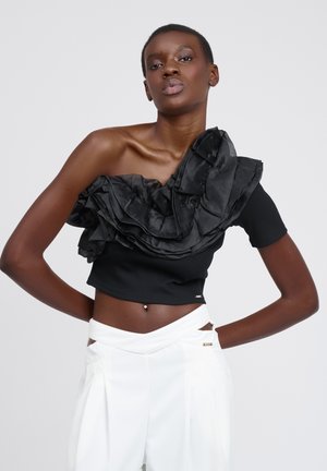 Kvinde med kort hår iført sort en-ærmet top med stor flæse og hvide høj-taljede bukser med unikt udskæringsdesign.