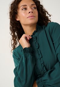 Teal blouse with ruffle details along the front, buttons, and gathered sleeves. Soft fabric with a smooth texture and relaxed fit.