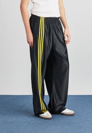 Person iført løse sorte bukser med tre lodrette gule striber og hvide sneakers, stående på blåt tæppe mod en ensfarvet baggrund.