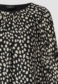 Schwarze Bluse mit plissiertem Design, geschmückt mit einem weißen abstrakten Muster. Der Stoff hat eine glatte Textur und einen Rundhalsausschnitt.
