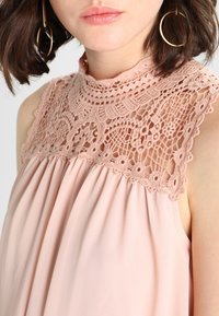 Débardeur couleur blush avec un empiècement en dentelle aux motifs complexes, tissu froncé en dessous, et un col montant pour une coupe décontractée.