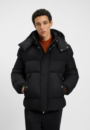 Giacca a vento nera con design imbottito, cappuccio staccabile e dettagli con bottoni a pressione. Presenta tasche laterali e un colletto alto.