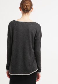 Femme avec les cheveux en chignon portant un pull à manches longues gris foncé avec des bordures gris clair, vue de dos sur un fond blanc uni.