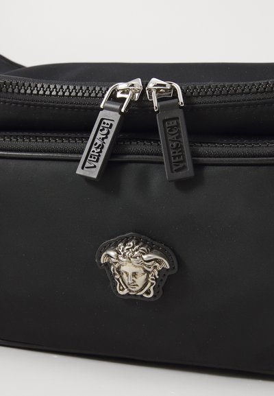 Črna najlon torba z dvema srebrnima zadrgama z zaponkami z logotipom Versace in osrednjim kovinskim emblemom Meduse. Teksture in elegantna zasnova.