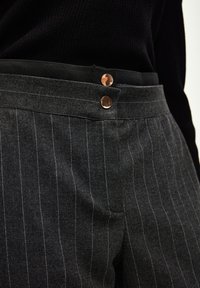 Pantalon gris foncé à fines rayures avec deux boutons métalliques à la taille, porté avec un haut noir côtelé.