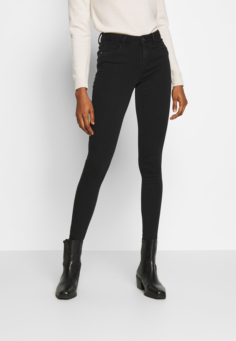 Personne portant un jean noir slim, des bottines noires et un haut blanc à manches longues, debout devant un fond blanc uni.